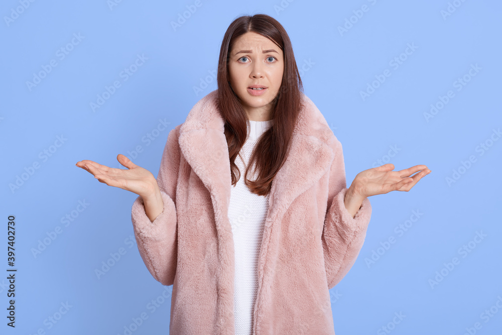 Insecure puzzled dark haired female has European appearance, spreads hands with doubt, dressed in pale pink fur coat, posing isolated over blue background. Insecure puzzled dark haired female has European appearance, spreads hands with doubt, dressed in pale pink fur coat, posing isolated over blue background.