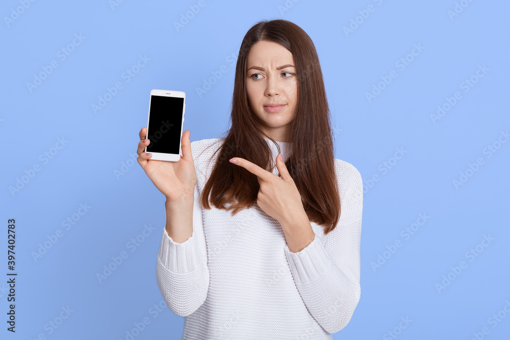 Winsome displeased woman has long brown hair, wears white sweater, points with index finger at smart phone with empty screen, demonstrates something strange, isolated over blue background.