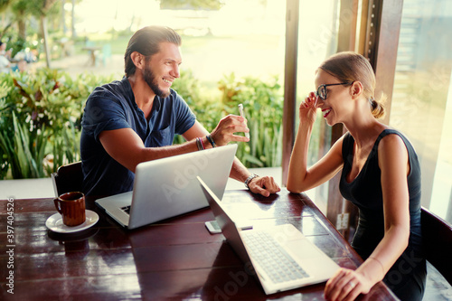 Coworking and freelance concept. Young bearded man showing smartphone to woman working together on laptop computer while sitting on cafe terrace.