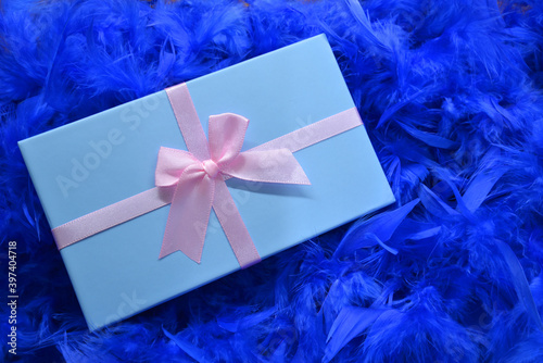 
Blue box with a gift on a blue feather background. Gift wrapped in gold with colored ribbons and blue feathers.