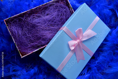 
Blue box with a gift on a blue feather background. Gift wrapped in gold with colored ribbons and blue feathers.