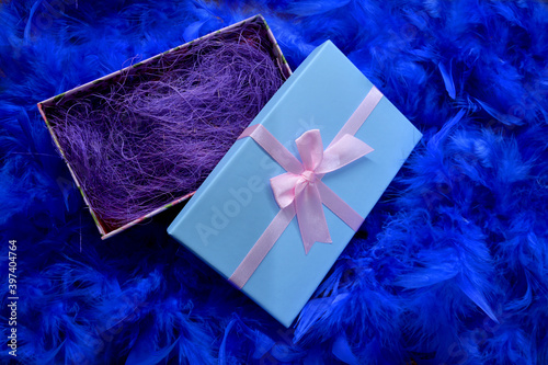 
Blue box with a gift on a blue feather background. Gift wrapped in gold with colored ribbons and blue feathers.