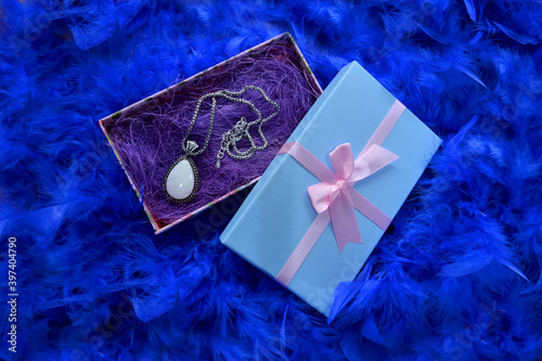 
Blue box with a gift on a blue feather background. Gift wrapped in gold with colored ribbons and blue feathers.