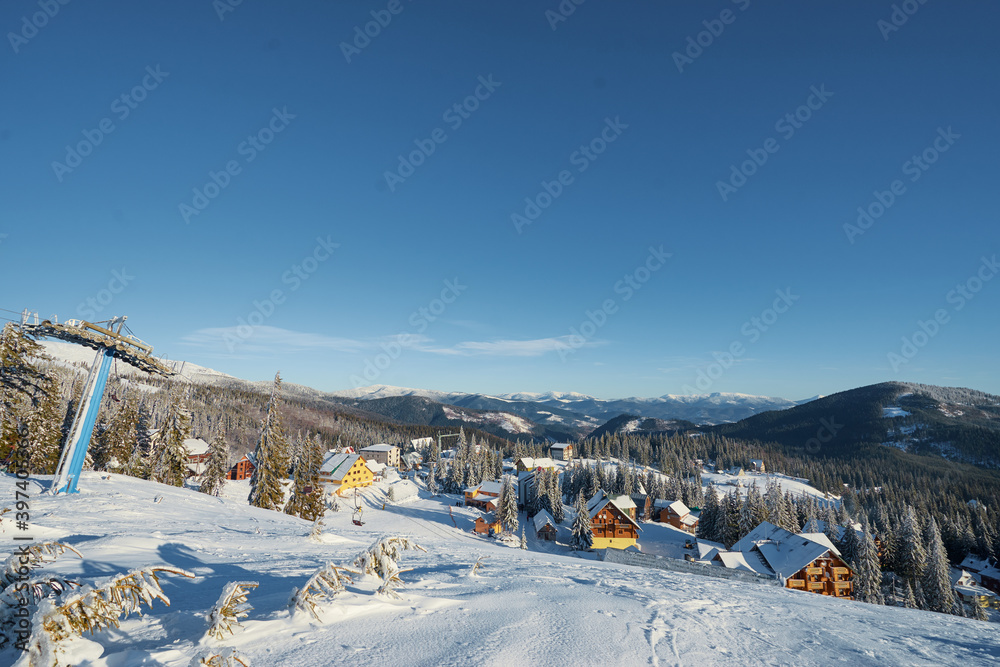 Beautiful white winter wonderland mountain scenery in the Carpathian with traditional houses. Snowy mountains and ski lifts. Skiers and snowboarders skiing downhill to village.