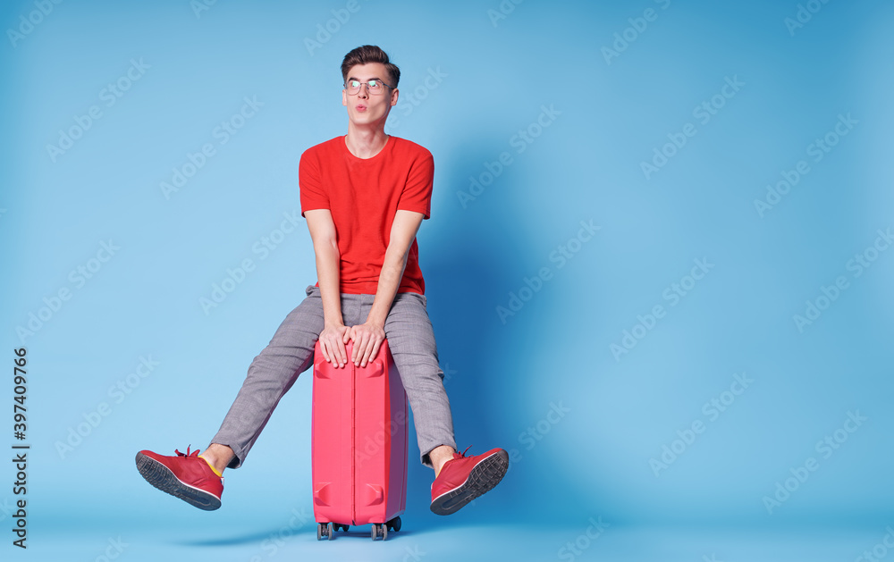 Travel concept. Full length colorful studio portrait of young man on valise on blue background.