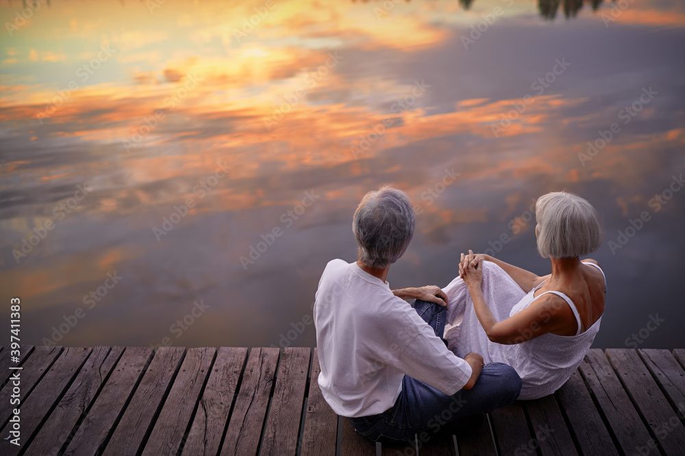 Romantic holiday. Senior loving couple sitting together on lake bank enjoying beautiful sunset. Romantic holiday. Senior loving couple sitting together on lake bank enjoying beautiful sunset.