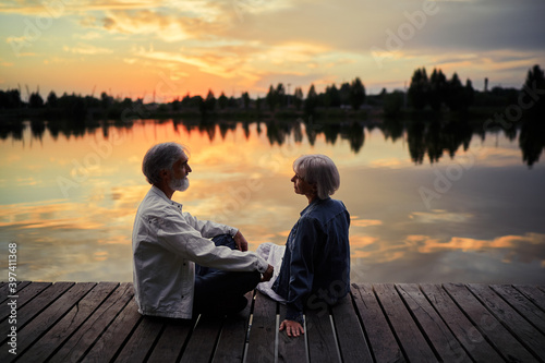 Romantic holiday. Senior loving couple sitting together on lake bank enjoying beautiful sunset.