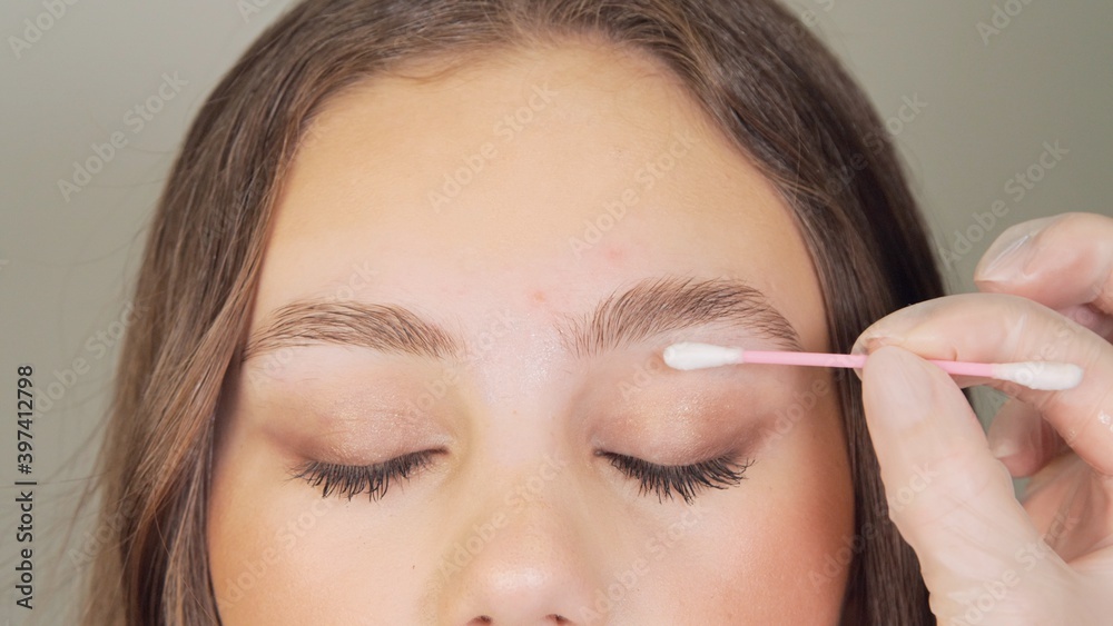 Cosmetologist wipes the client's eyebrows using cotton swabs. Beautician in gloves is doing permanent makeup to girl. Eyebrow correction. Preparation for lamination and coloring of eyebrows. Cosmetologist wipes the client's eyebrows using cotton swabs. Beautician in gloves is doing permanent makeup to girl. Eyebrow correction. Preparation for lamination and coloring of eyebrows.