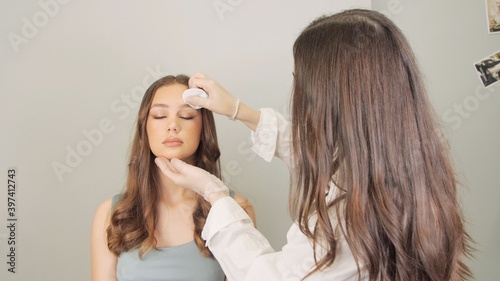 Cosmetologist wiping eyebrows using cotton pads. Beautician rubs client's face with a cotton pad. Beautician in gloves is doing permanent makeup to girl. Eyebrow correction.