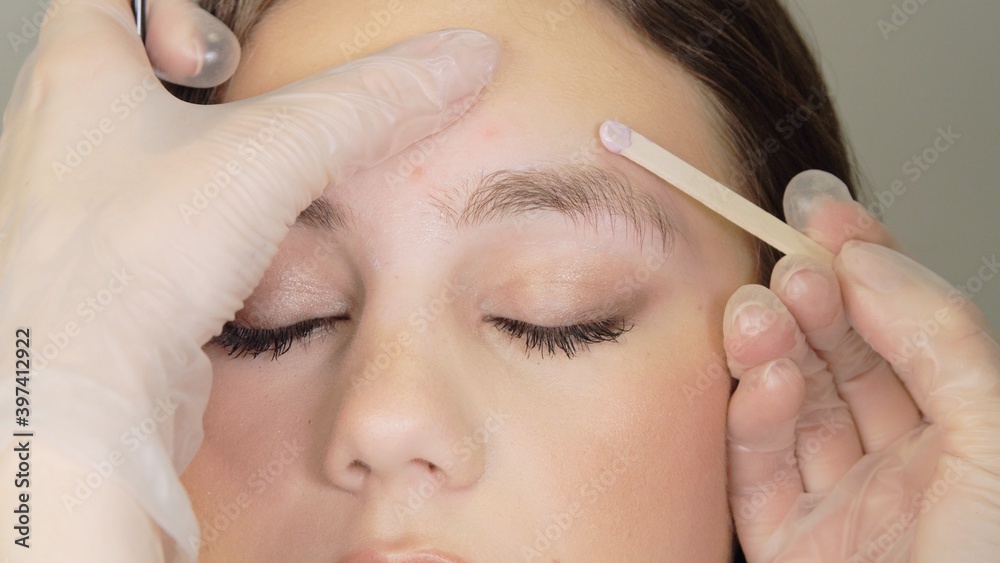 Female well-groomed hand in white medical gloves holds a wooden stick with hot wax for eyebrow shaping near female  beautiful face. Wax correction of the shape of the eyebrows with spatula. Close up.