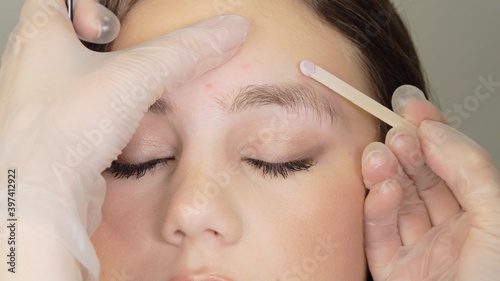 Female well-groomed hand in white medical gloves holds a wooden stick with hot wax for eyebrow shaping near female beautiful face. Wax correction of the shape of the eyebrows with spatula. Close up.
