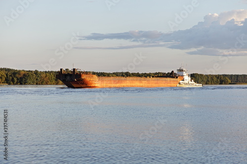 Soviet pusher cargo tug ship floating with empty barge on the Volga river on green forest shore and blue sky background, side view at summer day - river logistics transportation delivery by water  © Ilya