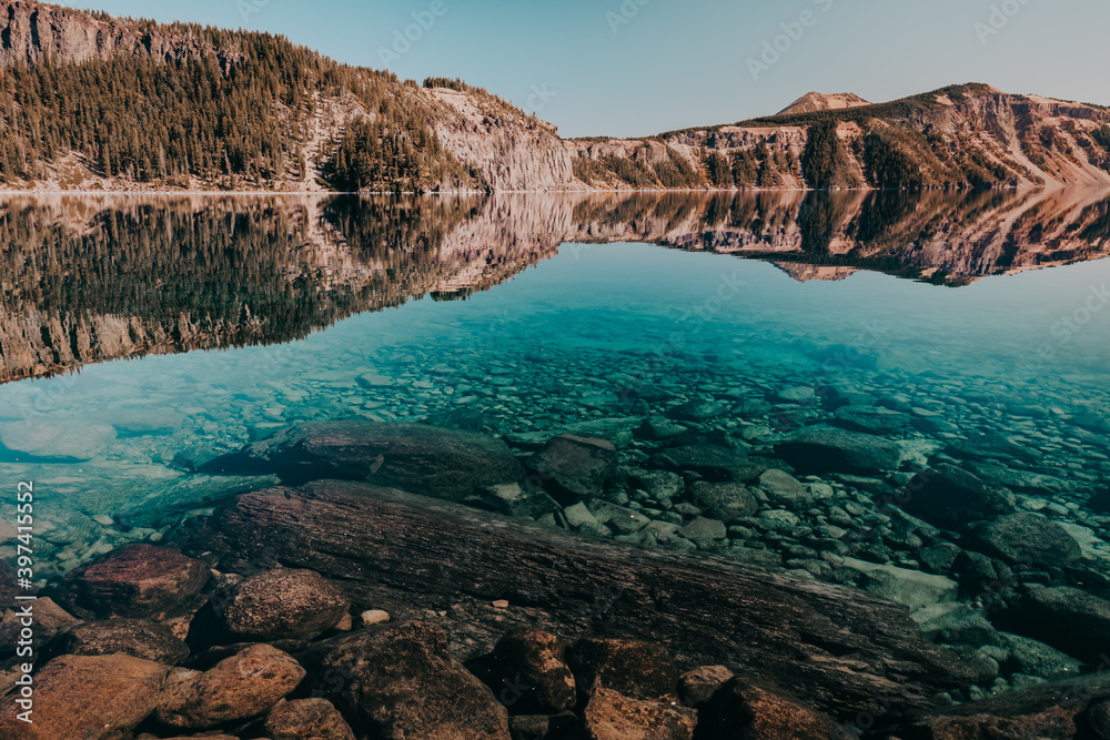 Reflections of Cleetwood Cove on Crater Lake at Crater Lake National Park, Oregon, USA. Crystal clear blue water with visible rocks and logs. 