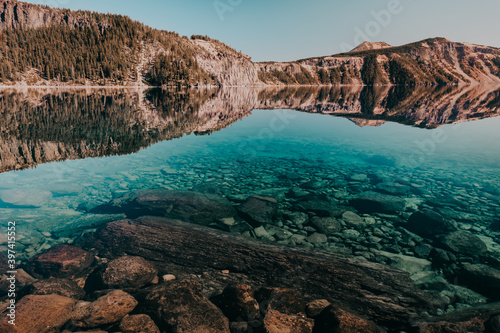 Reflections of Cleetwood Cove on Crater Lake at Crater Lake National Park, Oregon, USA. Crystal clear blue water with visible rocks and logs. 