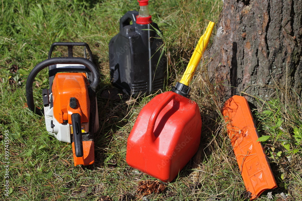 Outdoor chainsaw refueling - motorsaw, special plastic red can with gasoline fiel mix and chain oil canister, lumber sawing equipment near woon trunk at Sunny summer day on green grass background Outdoor chainsaw refueling - motorsaw, special plastic red can with gasoline fiel mix and chain oil canister, lumber sawing equipment near woon trunk at Sunny summer day on green grass background