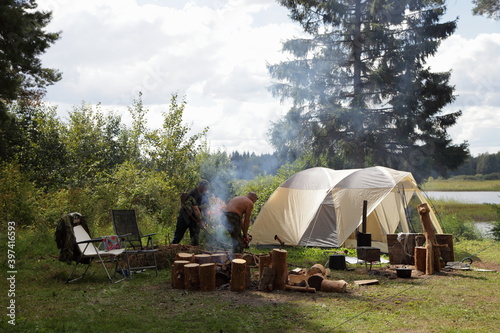 Tourist camp site tourist life, two scout men chopping firewood on camping tent and pine trees backhround on a Sunny summer day in an forest clearing on river shore, outdoor tourism romantic lifestyle