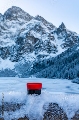 A red and black thermos cup with a hot drink (tea) placed in the frozen snow against the background of mountain peaks near the Morskie Oko Lake. Tatra Mountains, Poland. © gubernat