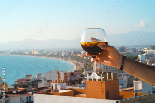 A hand raises a cup of an alcoholic cocktail froma roof top bar in a hotel at Mallorca with the bay of Playa de Palma a the background. Tourism, relax and luxury concept. photo