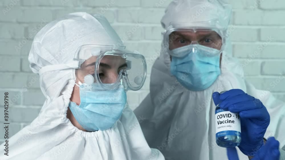 A scientist wearing a mask and protective suit passes a bottle of COVID-19 vaccine to his colleague. Container with blue liquid. Prevention, immunization and treatment from coronavirus infection 4k A scientist wearing a mask and protective suit passes a bottle of COVID-19 vaccine to his colleague. Container with blue liquid. Prevention, immunization and treatment from coronavirus infection 4k