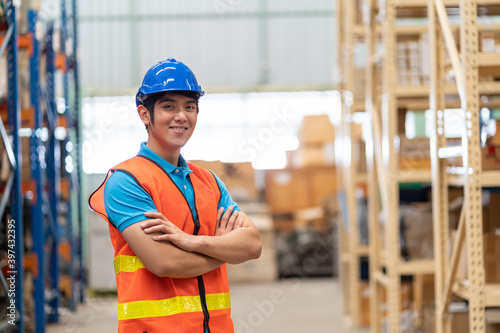 Confident smiling Asian male warehouse worker in safety vest and helmet standing with arms crossed in storage warehouse with shelf pallet spare parts parcel background