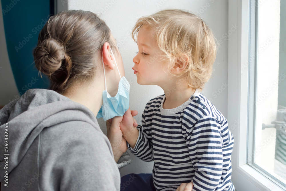 Child in home quarantine taking the medical mask off his mothers face and kissing her during coronavirus COVID-2019 and flu lock down. Selective focus Child in home quarantine taking the medical mask off his mothers face and kissing her during coronavirus COVID-2019 and flu lock down. Selective focus