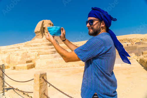 A young man putting on the mask facing the Great Sphinx of Giza and the Pyramids of Giza in the background  tourism in the middle of the coronavirus pandemic  covid-19. In the city of Cairo  Egypt