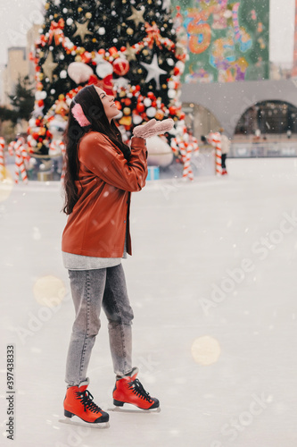 Happy carefree beautiful girl in stylish jacket, furry earmuffs catch snow on palm, smile on ice skating rink. Cheerful young pretty woman have fun, skate on outdoor ice rink. Christmas winter holiday