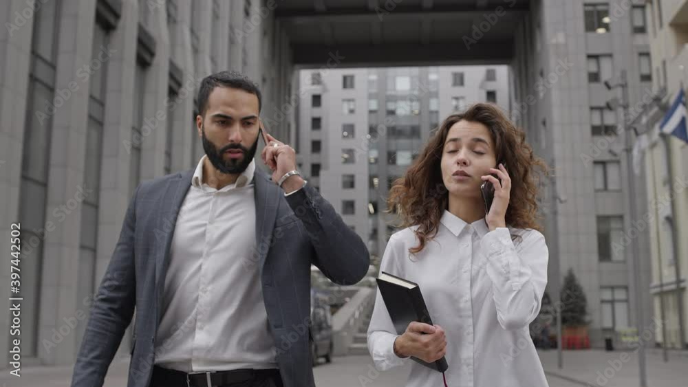 Two Business People Walk Down And Talk On The Phone. Young Man Of Oriental Nationality And Pretty Girl With Long Curly Hair. Two Business People Walk Down And Talk On The Phone. Young Man Of Oriental Nationality And Pretty Girl With Long Curly Hair.