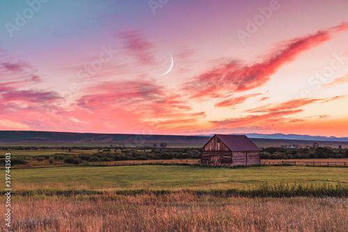 A beautiful sunset in a rural Montana scene. A simple barn sits in a field as the sun sets while the crescent moon rises above. photo