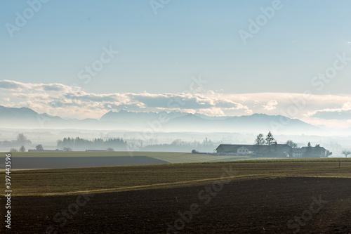 Alpen Panorama im Chiemgau in Bayern bei Föhn im Winter mit leichtem Nebel , blauem Himmel Wolken und Sonnenschein mit Häuser, Bäumen und Bergen