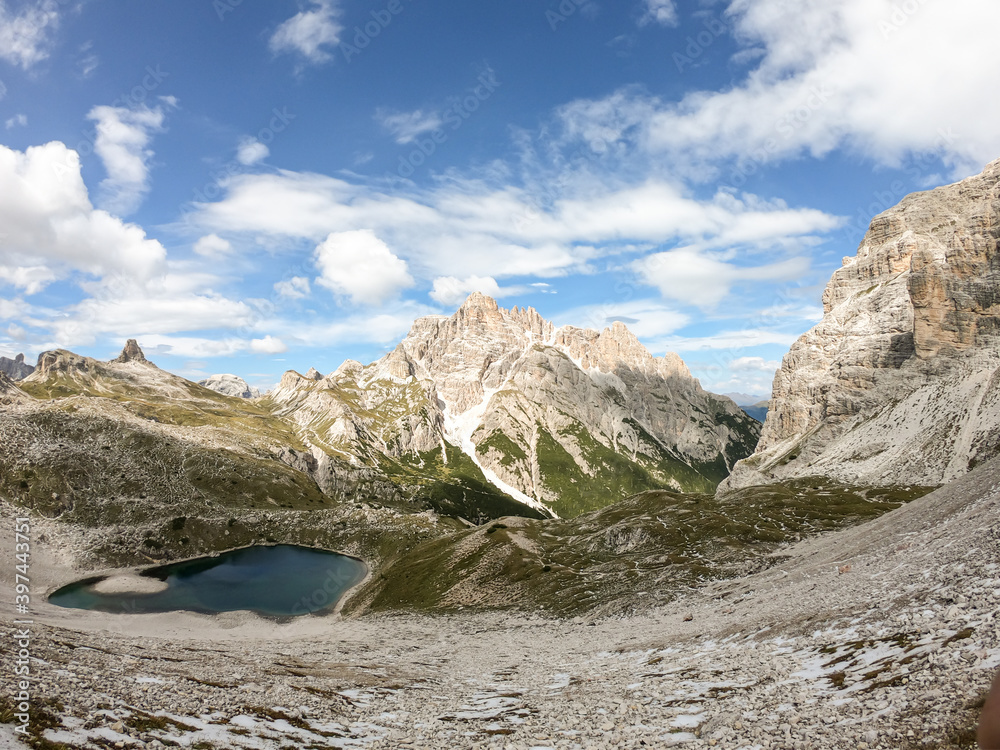Obraz premium A panoramic view on Dolomites in Italy. There are sharp and steep mountain slopes around. At the bottom of a small valley there is a small navy blue lake. The sky is full of soft clouds. Raw landscape