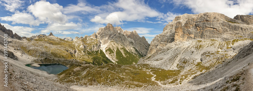 A panoramic view on Dolomites in Italy. There are sharp and steep mountain slopes around. At the bottom of a small valley there is a small navy blue lake. The sky is full of soft clouds. Raw landscape © Chris