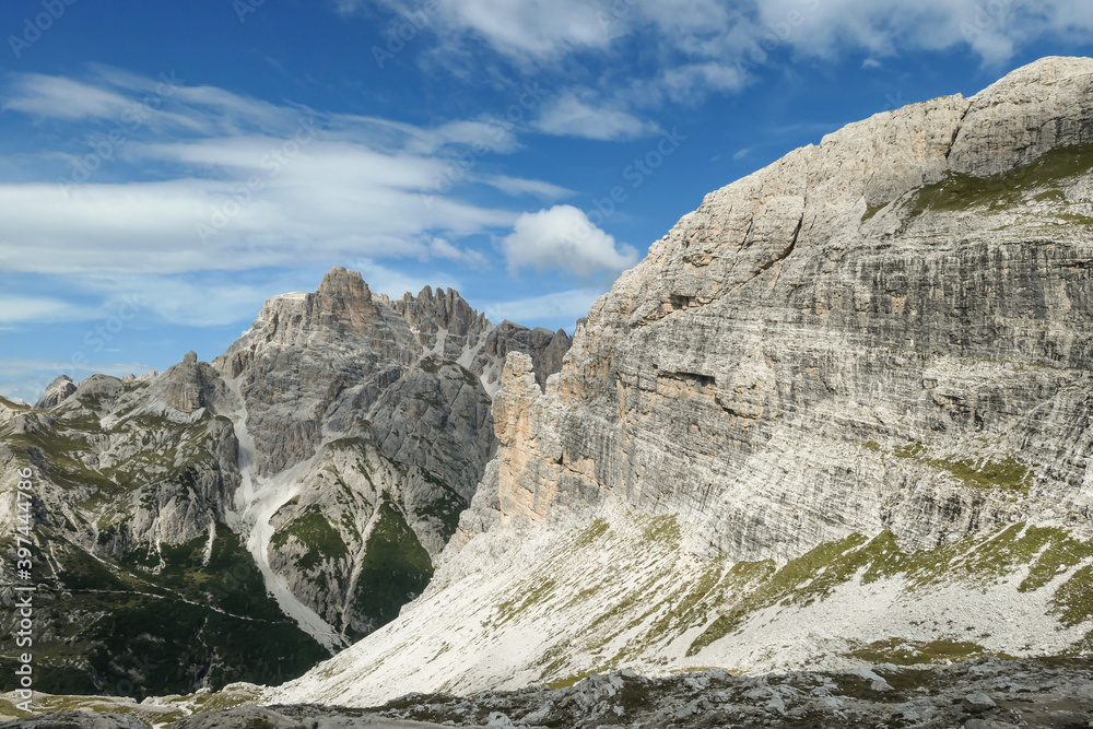 A panoramic view on Dolomites in Italy. There are sharp and steep mountain slopes around. Lots  of lose stones and pebbles. The sky is full of soft clouds. Raw landscape. Serenity and calmness