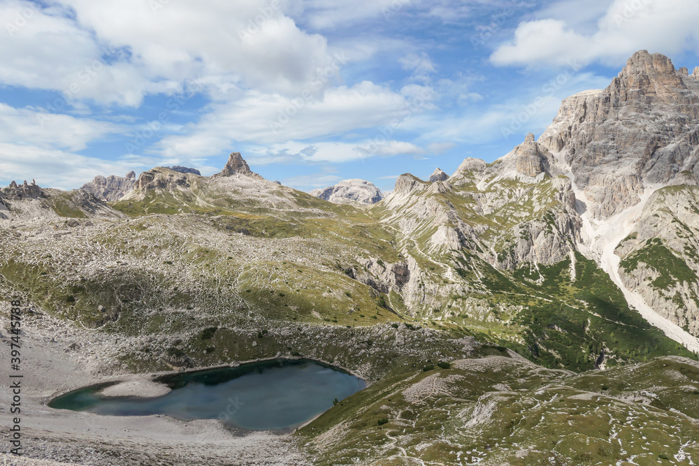 A small, navy blue lake at the bottom of the valley in Italian Alps. The lake is surrounded by high and steep peaks. The slopes are lush green. The sky is full of soft clouds. Raw landscape. Remedy
