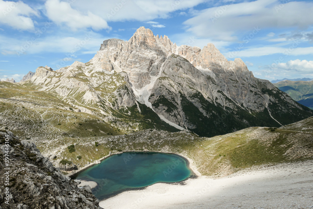 Obraz premium A small, navy blue lake at the bottom of the valley in Italian Alps. The lake is surrounded by high and steep peaks. The slopes are lush green. The sky is full of soft clouds. Raw landscape. Remedy
