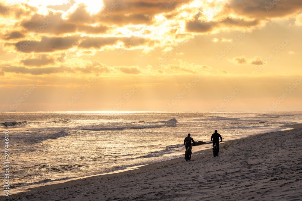 A pair of people cycling down the beach on fat bikes at sunset. 