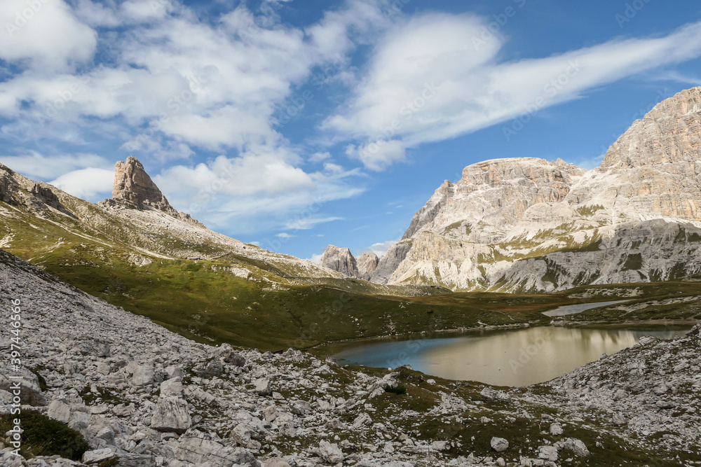 Obraz premium A small, navy blue lake at the bottom of the valley in Italian Alps. The lake is surrounded by high and steep peaks. The slopes are lush green. The sky is full of soft clouds. Raw landscape. Remedy