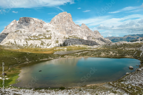 A small  navy blue lake at the bottom of the valley in Italian Alps. The lake is surrounded by high and steep peaks. The slopes are lush green. The sky is full of soft clouds. Raw landscape. Remedy