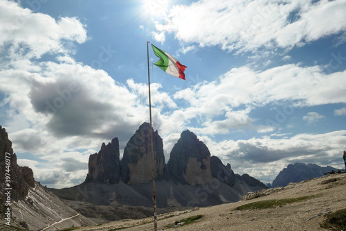 An Italian flag on a high pole waving above the high and sharp peaks of Dolomites in Italy. The sky is full of soft clouds. Raw and desolated landscape. Serenity and calmness. Conquering a territory