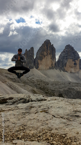Man in hiking outfit jumping in front of the famous Tre Cime di Lavaredo (Drei Zinnen), mountains in Italian Dolomites. Desolated and raw landscape, full of lose stones. Overcast. Achievement and fun