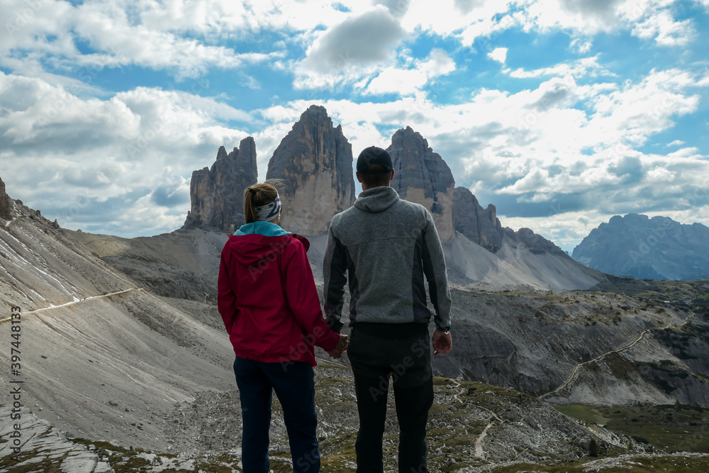 A couple in hiking outfit holding hands and enjoying the view on the famous Tre Cime di Lavaredo (Drei Zinnen) in Italian Dolomites. Desolated and raw landscape, full of lose stones. Overcast. Love