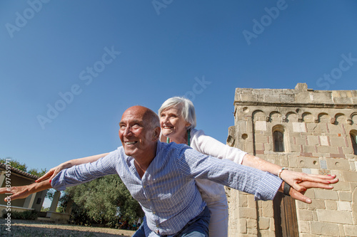 Coppia di anziani felici che giocano simulando l'aeroplano vicino aa una chiesa in un parco all'aria aperta. 