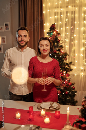 Happy couple with champagne standing by fesive table against Christmas tree