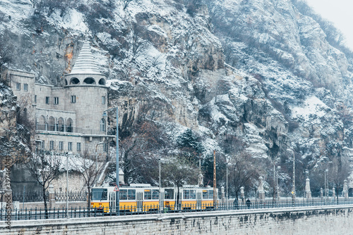 Yellow tram with Gellert hill covered with snow on background during cloudy winter day