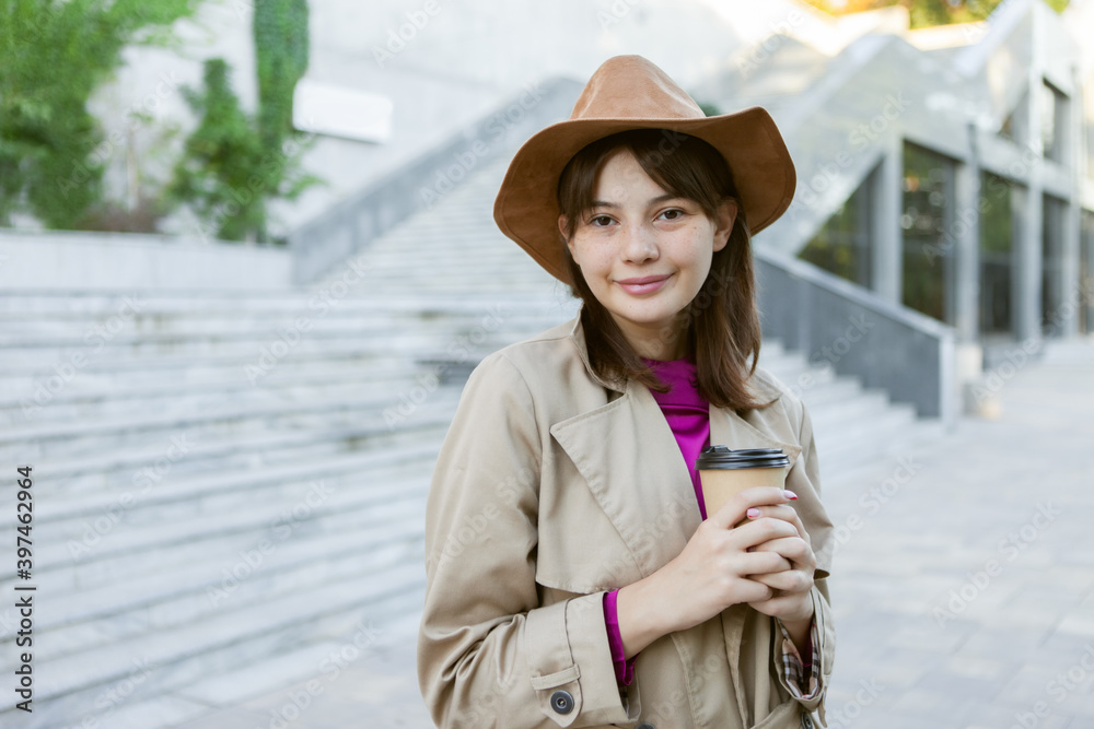 Fashion young woman in a felt hat and an autumn trench coat holding a cup of coffee in a city park.