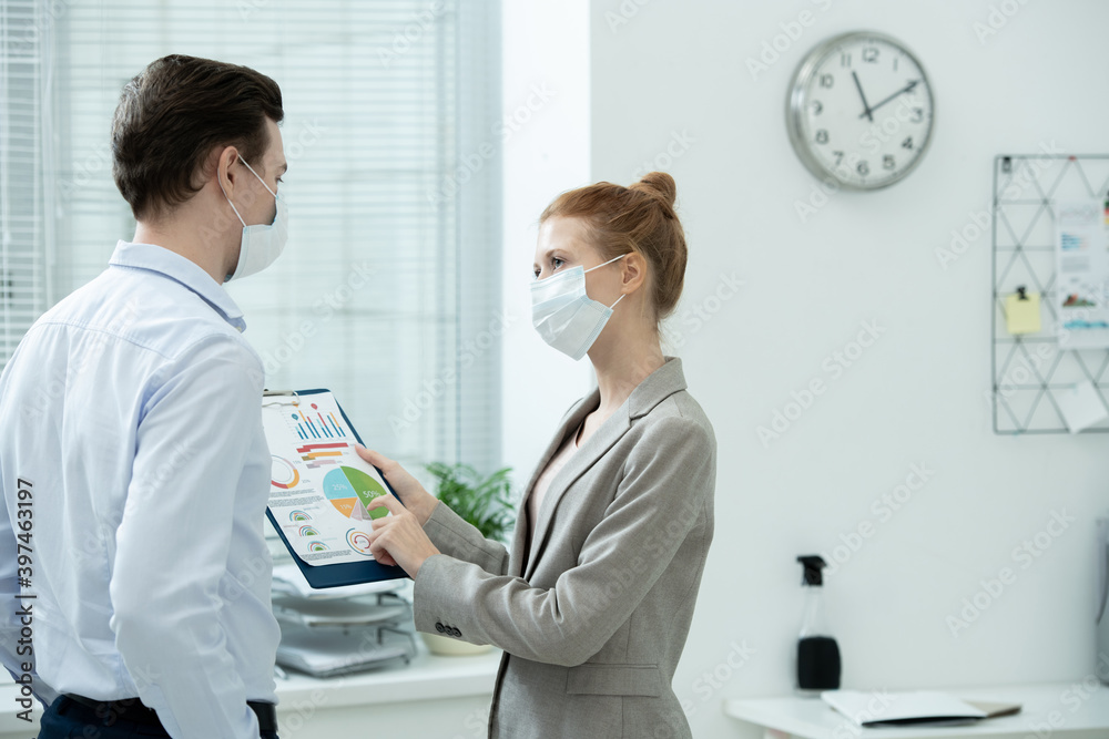 Two young elegant accountants in protective masks discussing financial diagram