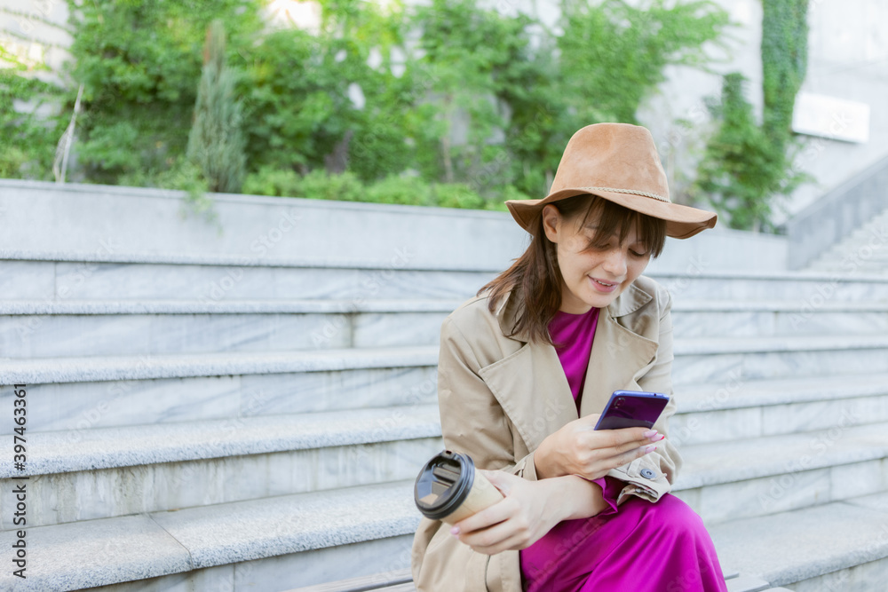 Obraz premium Young fashion woman dressed in autumn clothes sits on bench and uses a smartphone outdoors