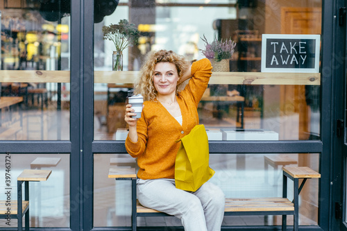 Blond curly female customer in yellow knitted cardigan, sitting outdoor near glass window of store or zero waste shop, on bench, drinking coffee from reusable silicone cup at warm aututmn day. photo