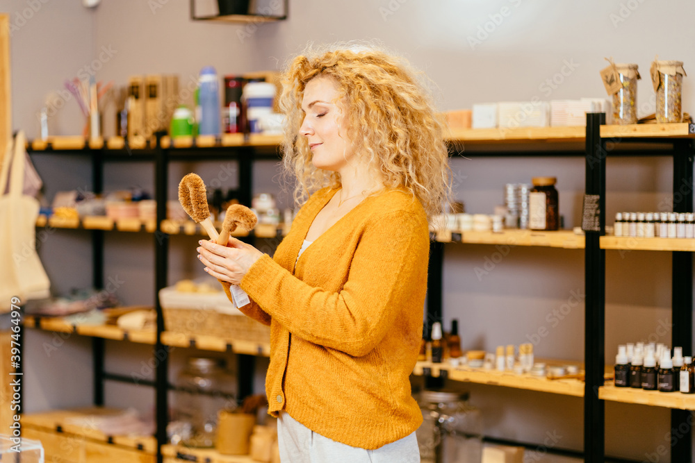 Vivacious blond curly woman on yellow background holds dishwashing brushes made from coconut flakes with bamboo handle. Biodegradable kitchen accessories. Zero waste and no plastic concept.