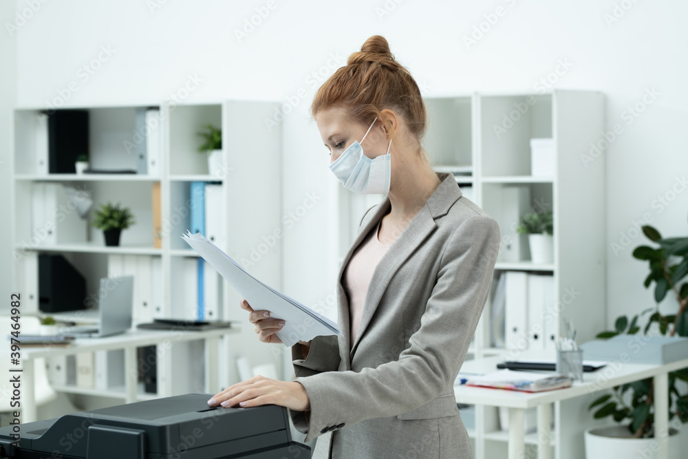 Young businesswoman in protective mask looking at paper by xerox machine